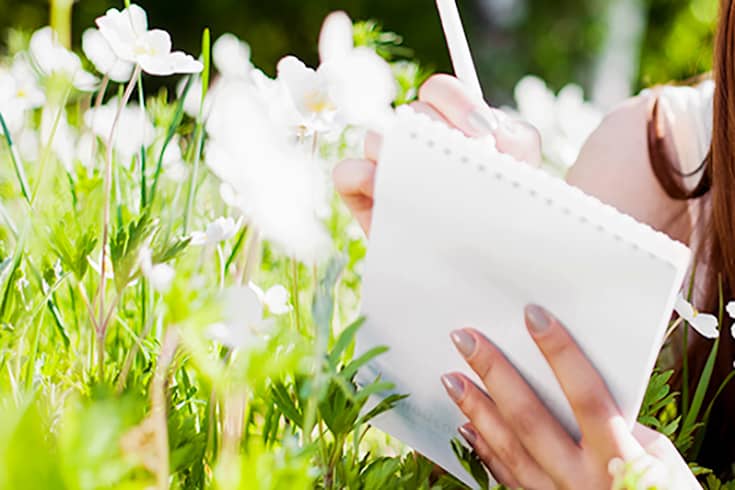 Woman in a garden of white flowers, taking notes in a notebook.